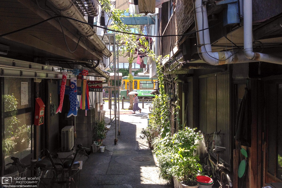 Through an alley of residential buildings in Kagoshima on the south western tip of the island of Kyushu in Japan, I captured this view of a city tram and a pedestrian carrying a parasol.