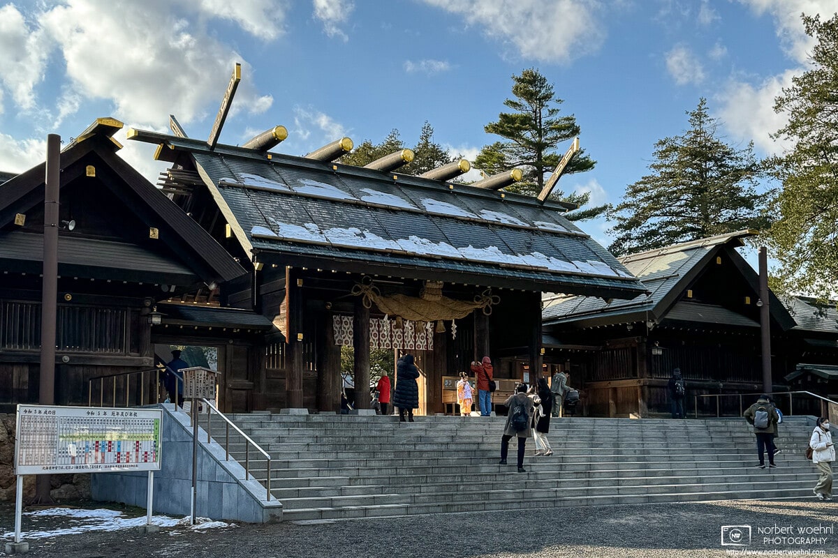 Outside the Main Gate of Hokkaido Jingu, a shrine in Sapporo, Japan, we can see parents taking a photo of a girl wearing a colorful kimono. Other visitors are seen entering or leaving the shrine.