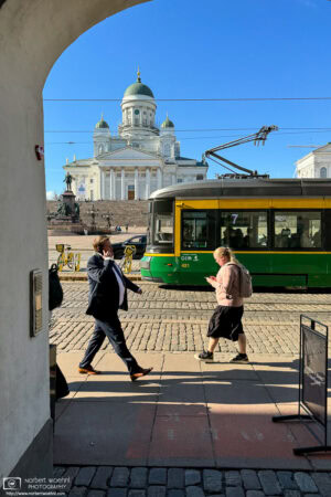 A street scene in front of Helsinki Cathedral. We can see a man in suit talking on his mobile phone, a woman with a backpack looking at her phone, and a green tram passing behind them, all in front of the white cathedral building in the background.
